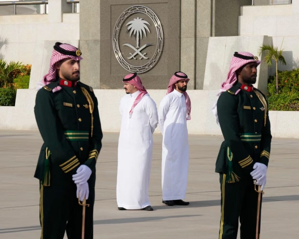 Two guards in green ceremonial uniforms stand at attention on either side of two men in white thobes in a plaza, with a palm-and-swords emblem on the wall behind them.