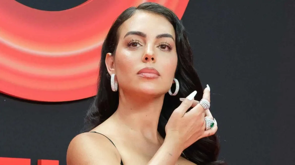Close-up of a woman with dark hair, bold makeup, large hoop earrings, and multiple rings, touching her hair against a red backdrop.