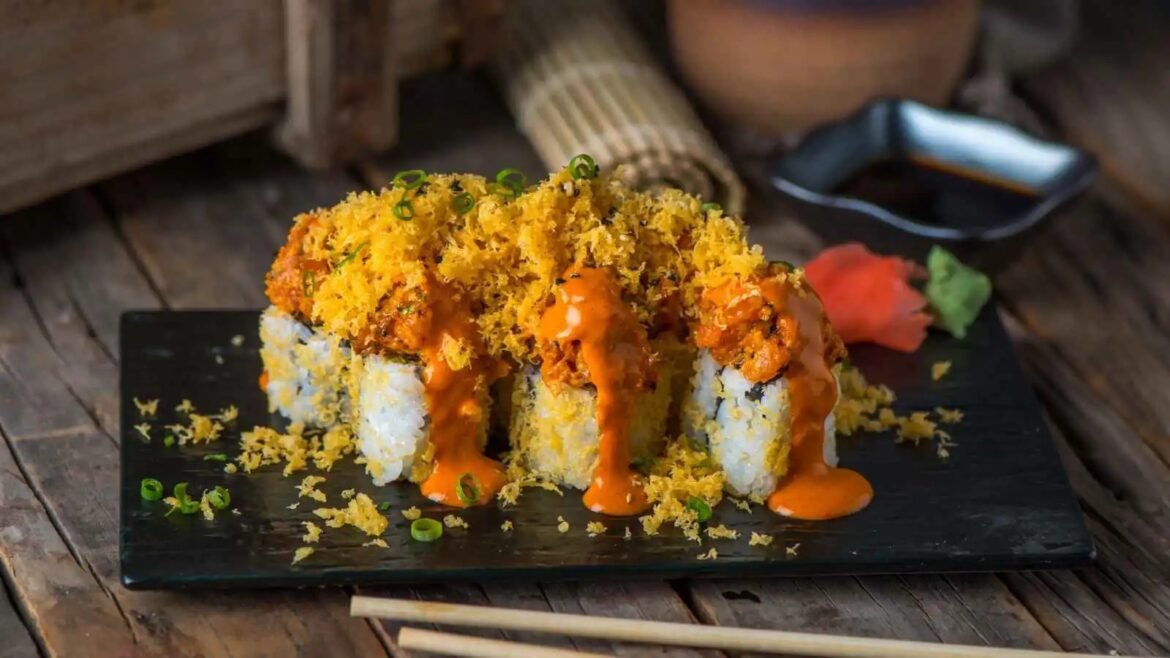 Sushi rolls topped with crispy tempura flakes and orange spicy sauce, garnished with green onions on a dark plate