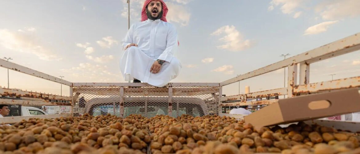Man in a white thobe and red keffiyeh sits cross‑legged on a rusted truck filled with dates, outdoors under a cloudy sky.