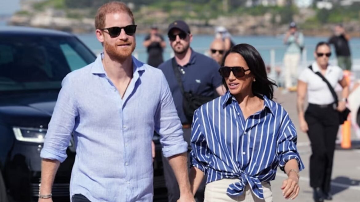 A man in a light blue shirt and a woman in a blue striped blouse walk together on a sunny waterfront boardwalk, both wearing sunglasses, with bystanders and a dark SUV in the background.