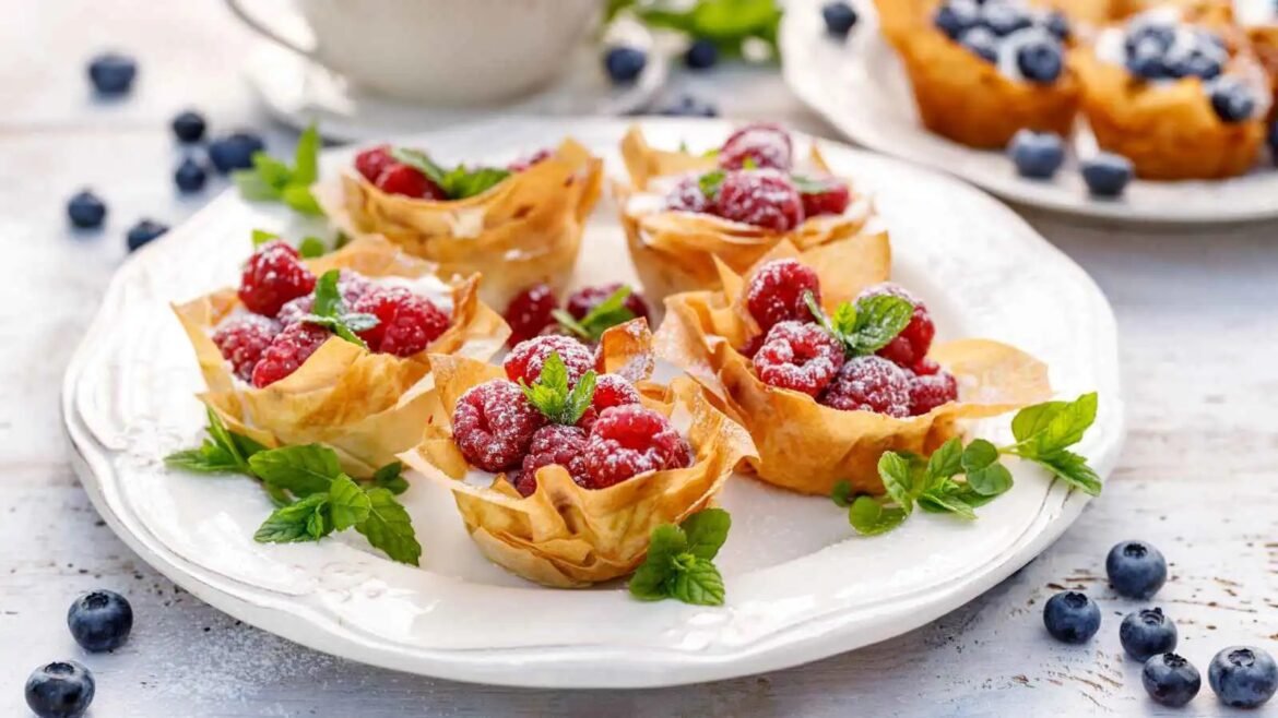 Phyllo cups filled with raspberries and mint on a white plate, dotted with blueberries nearby.
