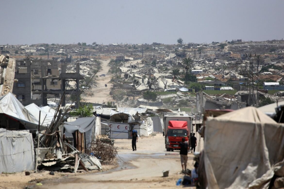 Dusty street through a tent-filled refugee camp with rubble and ruined buildings in the distance; a red truck is parked on the road.