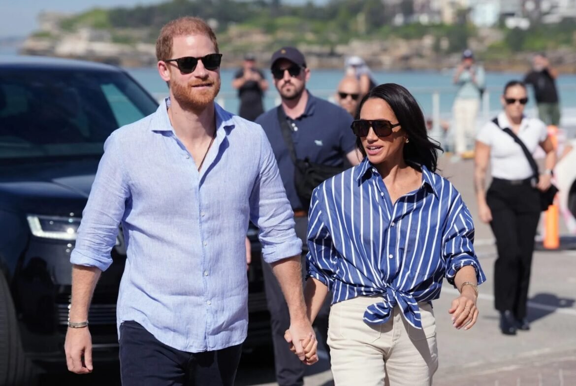 Couple wearing sunglasses walking hand in hand along a sunny waterfront promenade with onlookers in the background.
