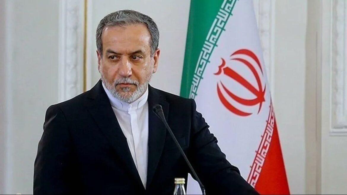 Middle-aged man in a dark suit speaks at a podium, with the Iranian flag in the background.
