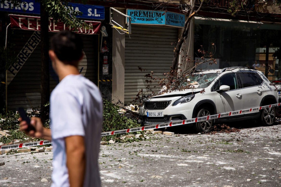 White SUV damaged by a fallen tree branch on a city street, debris scattered around and caution tape cordoning the area.
