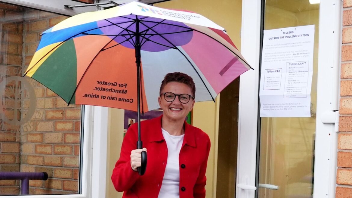 Smiling woman wearing a red coat and glasses holding a large rainbow-colored umbrella outside a brick storefront entrance.