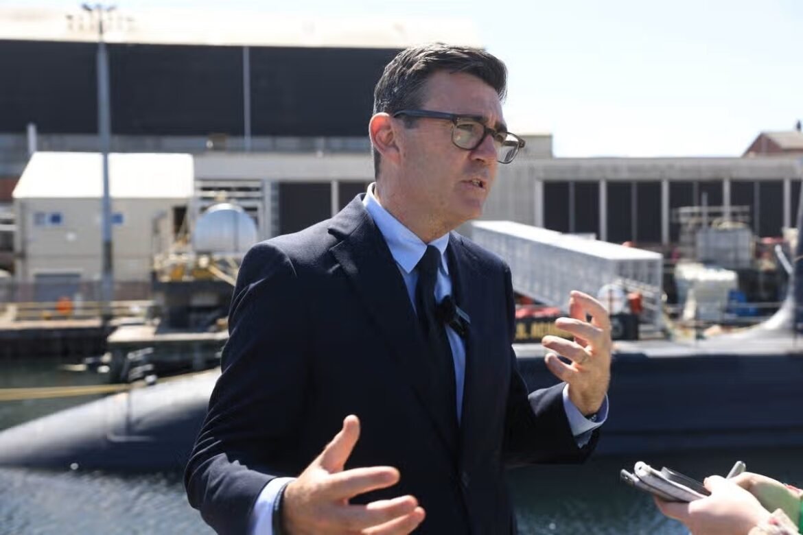Man in a navy suit and glasses speaks during an outdoor interview by a harbor.
