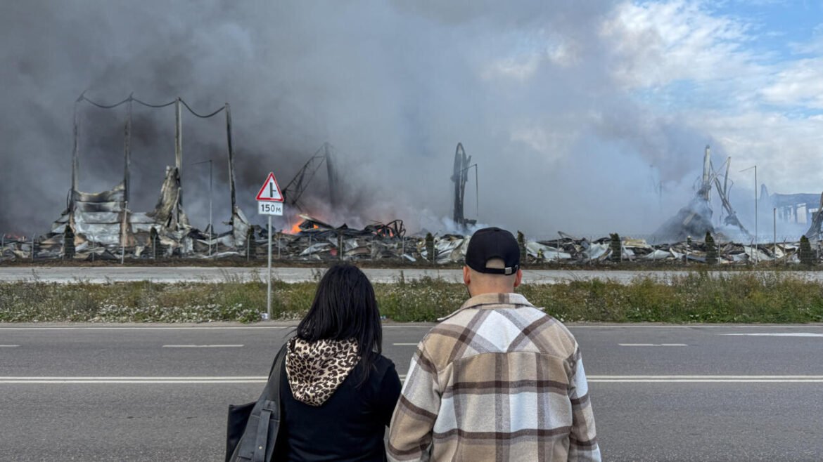 Two people stand on a highway shoulder, facing a large, smoky fire consuming a destroyed industrial site with charred frames and flames in the distance. Smoke fills the air as they look on.