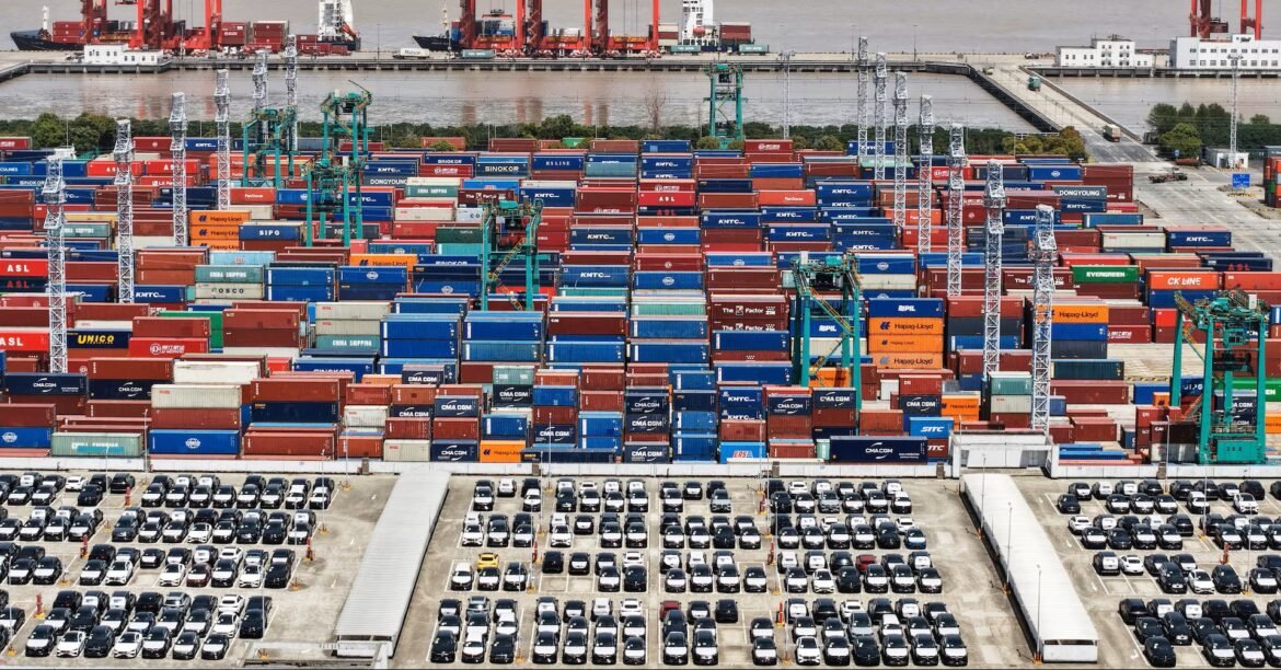 A busy container port with rows of stacked colorful shipping containers, tall cranes, and a foreground lot of parked cars.