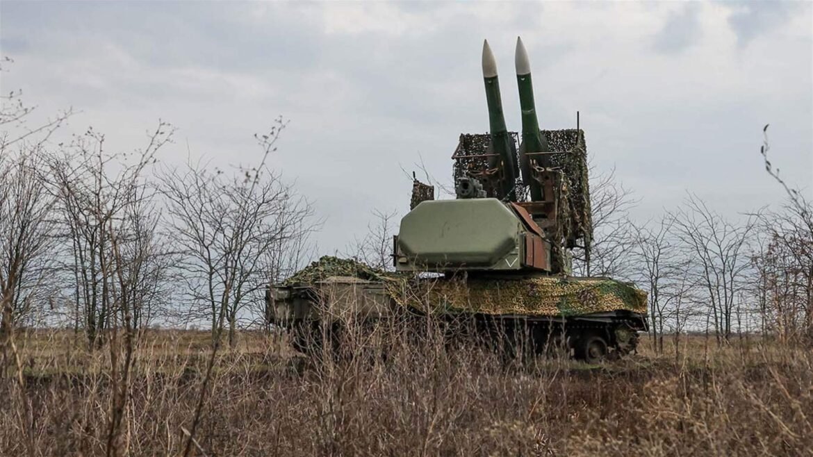 Tank with camouflage netting and two vertical missile tubes sits in a barren field among leafless trees under an overcast sky, during late fall/early winter.