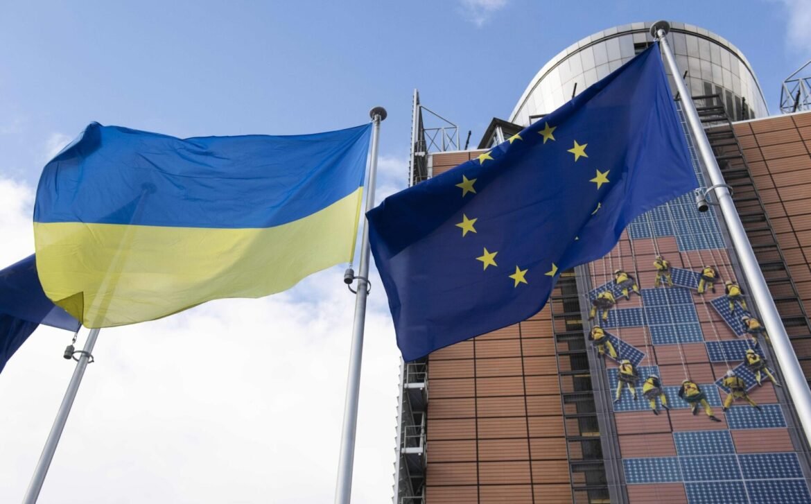 Ukraine and EU flags fly side by side in front of a modern solar-powered building.