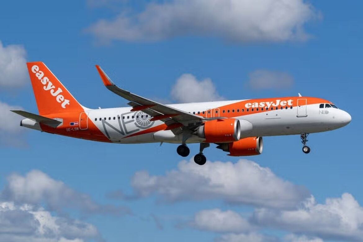 Orange and white easyJet passenger plane in midflight, landing gear down, against a blue sky with clouds.
