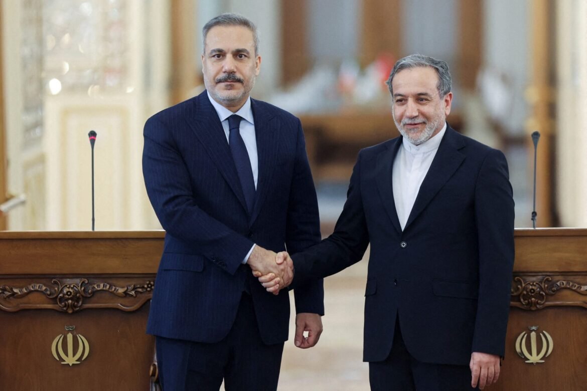 Two men in dark suits shake hands behind a carved wooden podium displaying the Iranian emblem in a formal setting.
