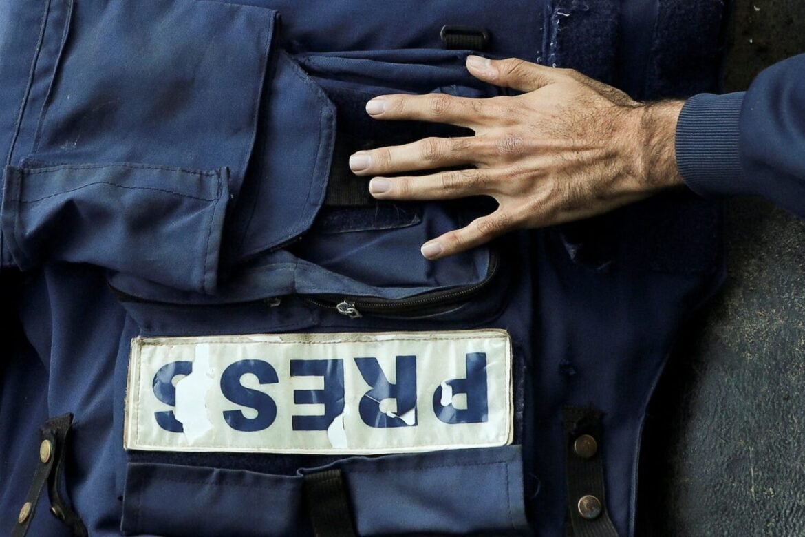 Navy-blue press vest with a worn 'PRESS' badge visible, a hand resting on the chest area.