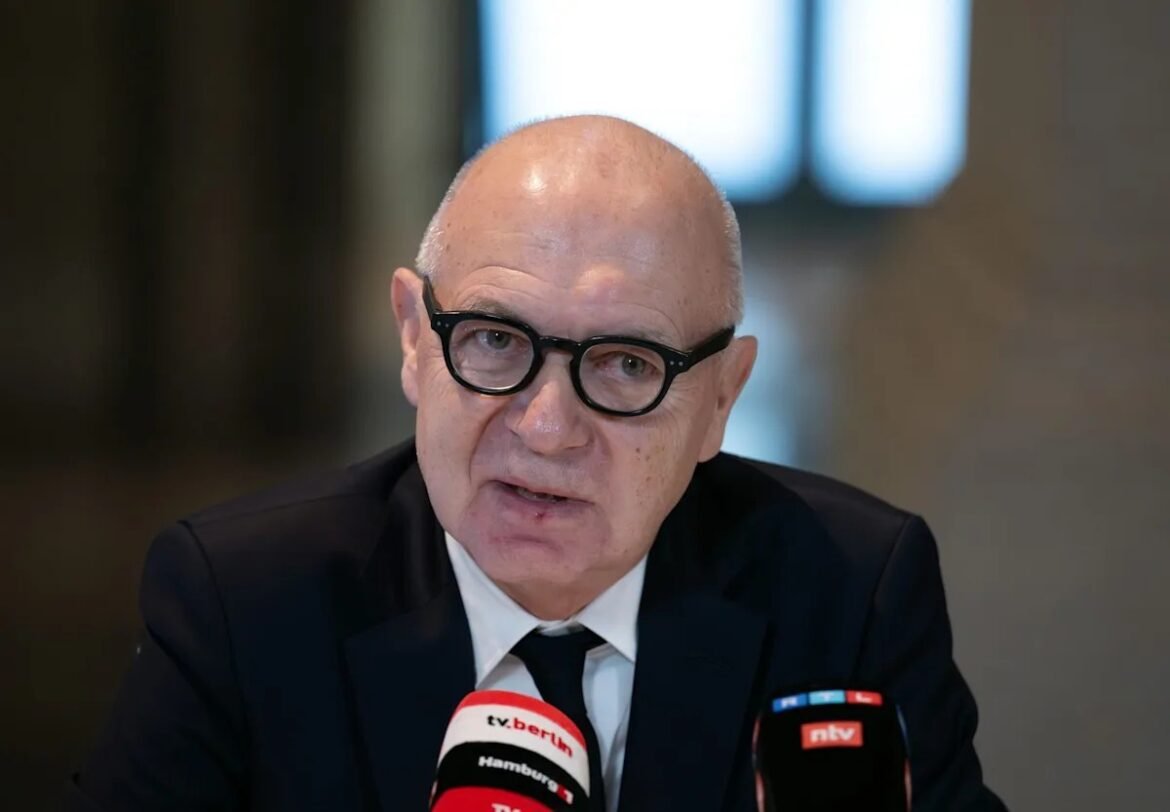 Older man in a dark suit and glasses speaks at a press conference, with two microphones labeled tv.berlin and ntv in front.