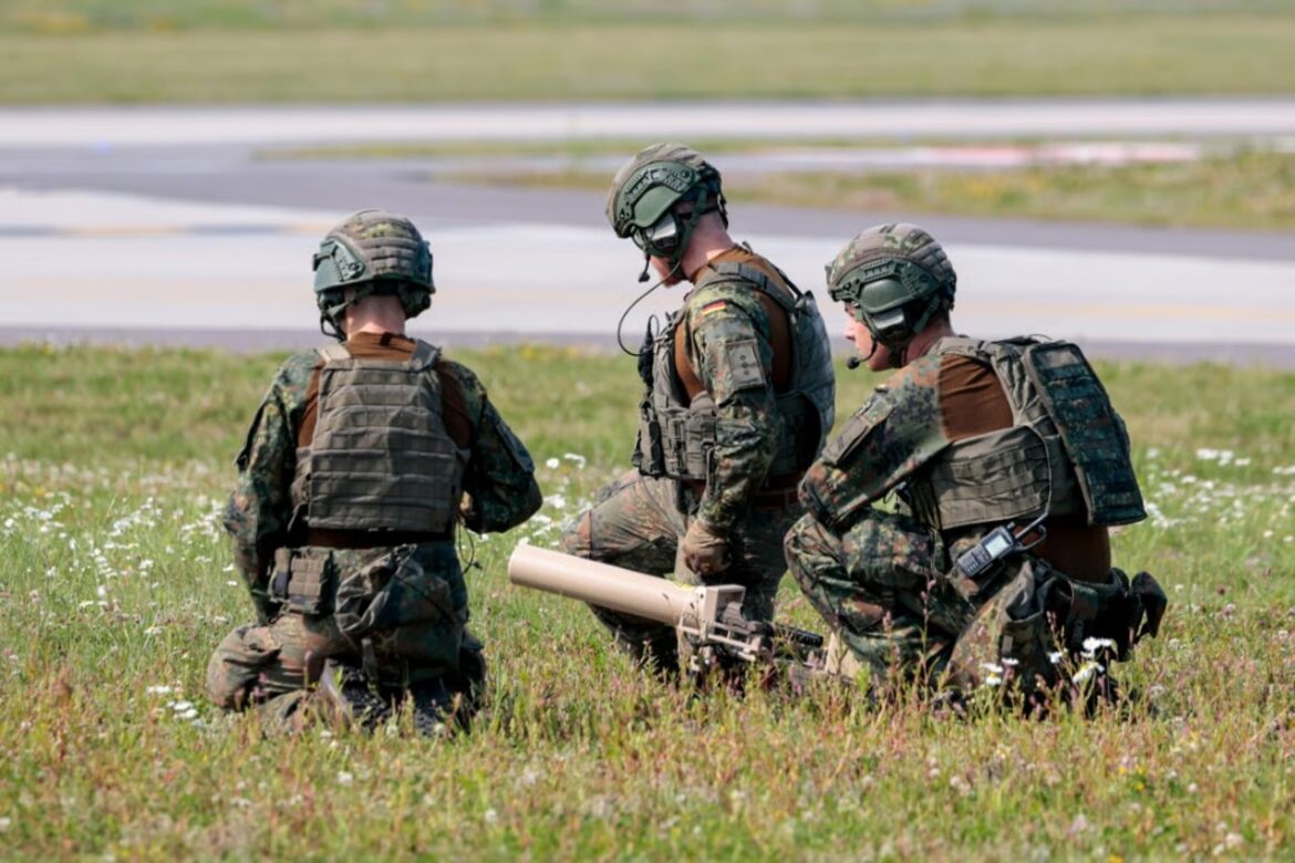 Three soldiers in camouflage kneeling in a grassy field near a runway, with a shoulder-fired launcher resting on the ground.
