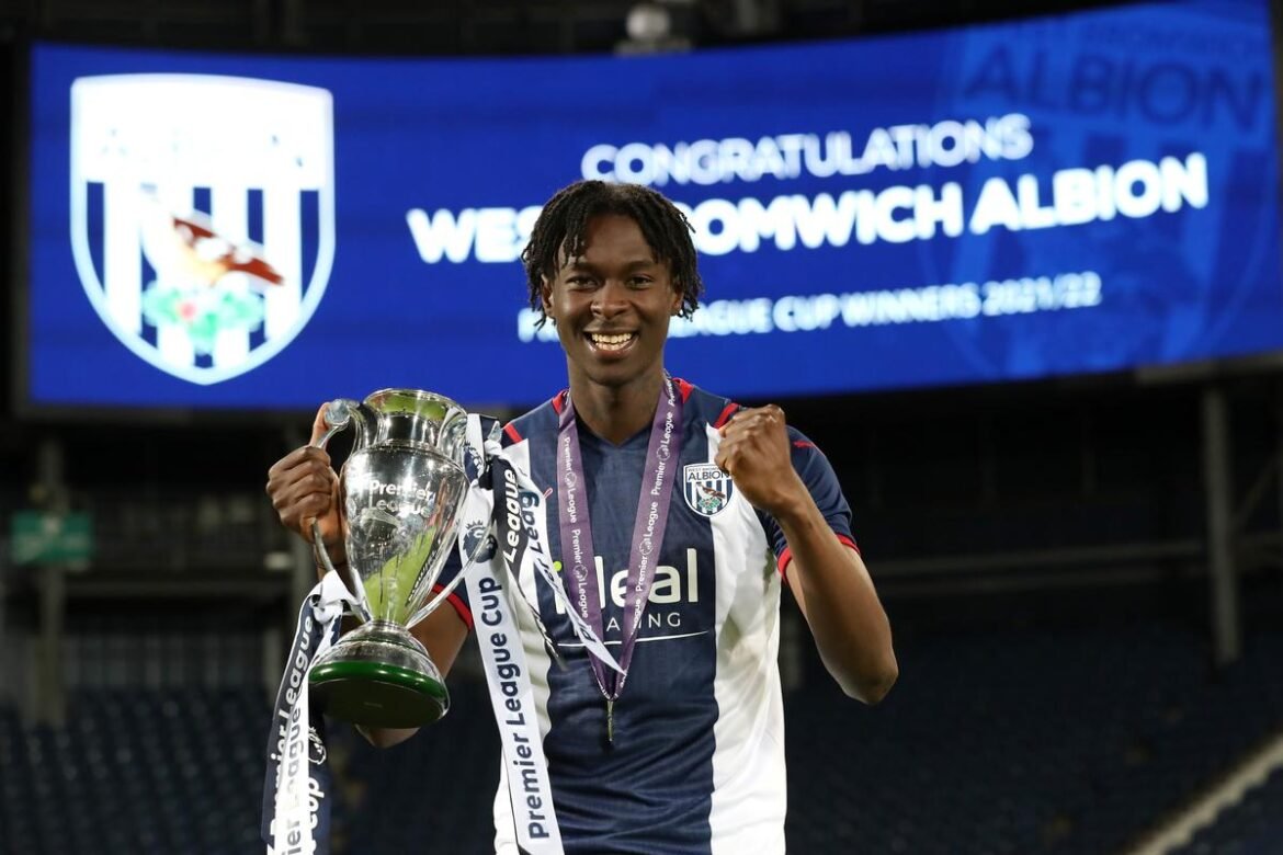 Young footballer in West Bromwich Albion kit smiles while holding a large silver trophy with ribbons at the stadium after a win, medals around his neck.