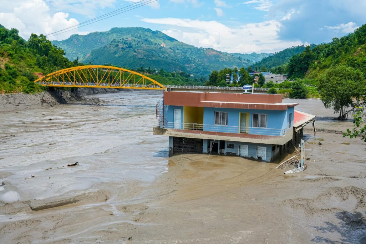 Blue and orange two-story house partially flooded by a muddy river with green hills and a yellow arch bridge in the background