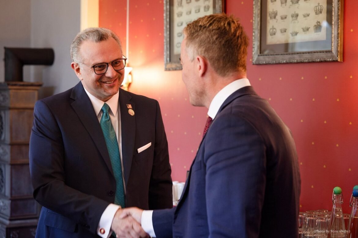 Two suited men shake hands and smile in a formal setting with red wallpaper and framed art behind them.