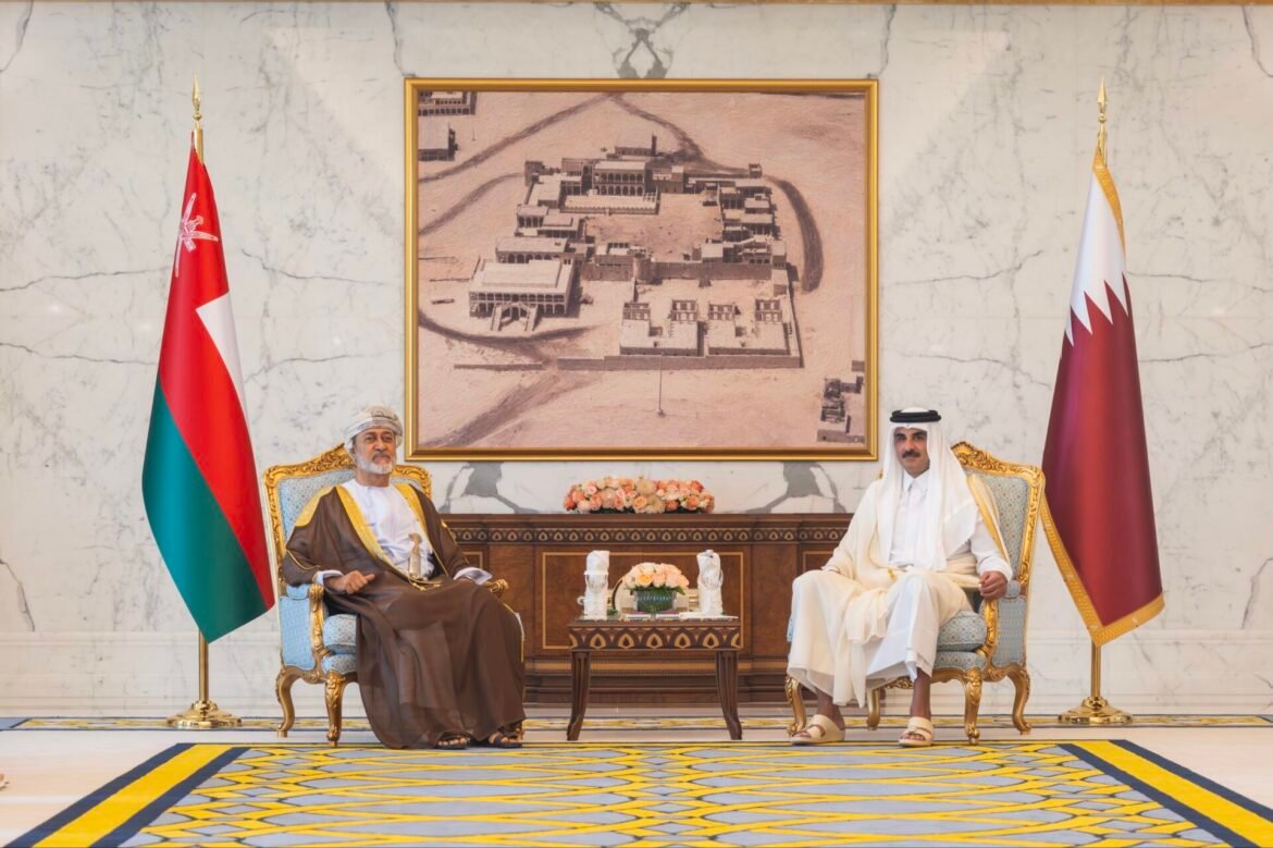 Two dignitaries in traditional attire sit on ornate gold chairs in a marble room, with Oman and Qatar flags beside them and a framed sepia map behind them.