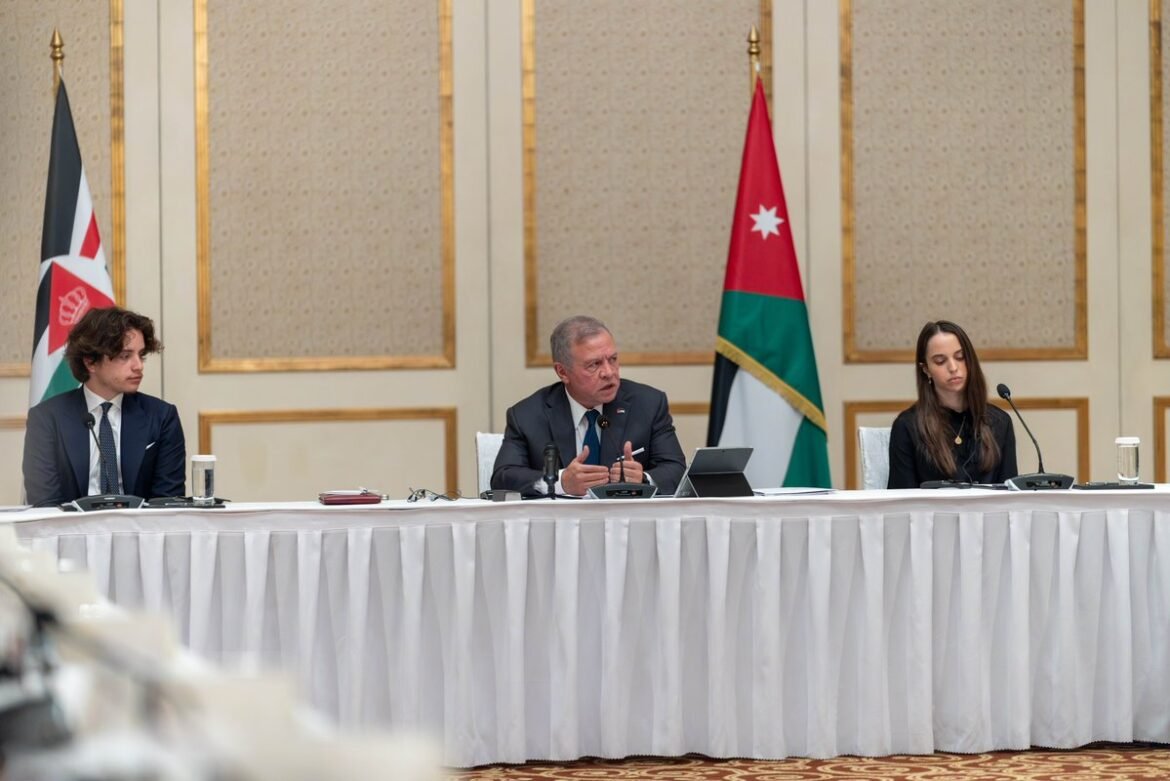 Three panelists seated at a long white-draped table in a formal meeting room, with Jordanian flags behind them