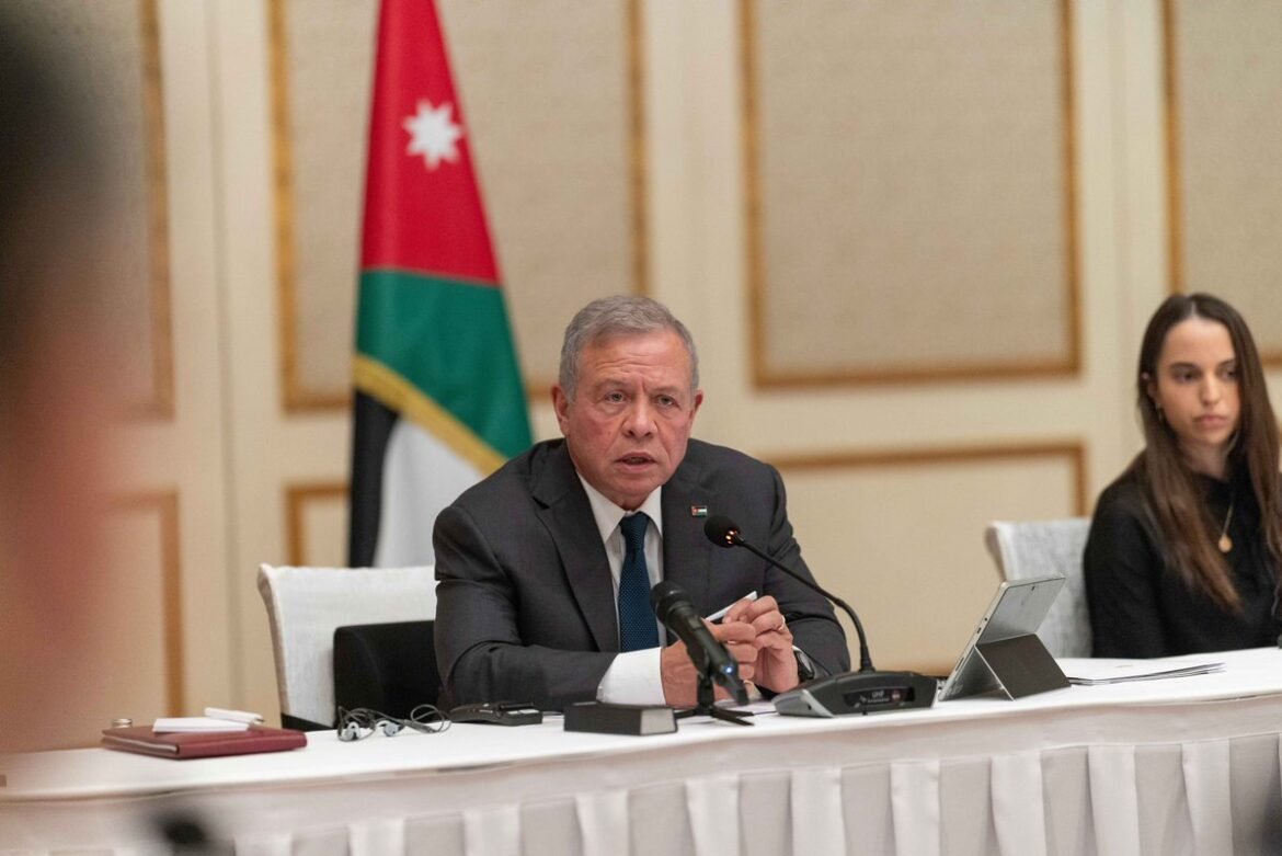 Middle-aged man in a suit speaks at a panel with a microphone, Jordan flag in the background.