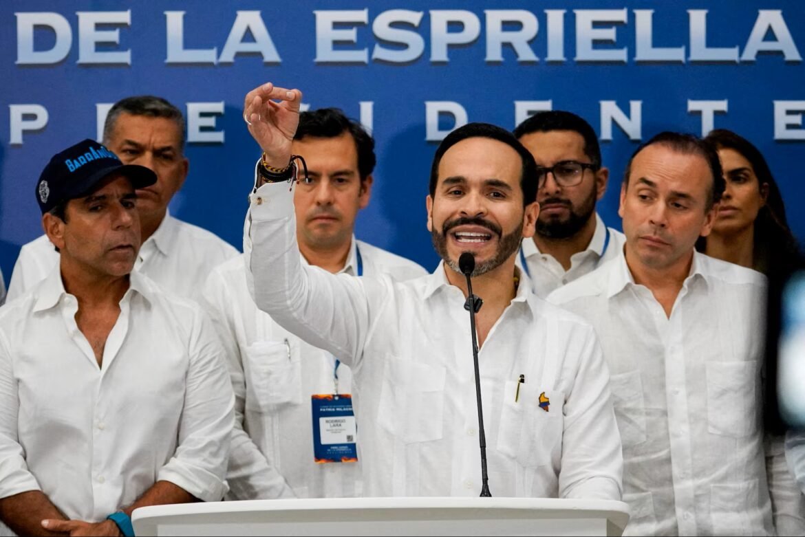 Man at a podium speaking into a microphone, raising his hand, with supporters in white shirts behind him at a political event.