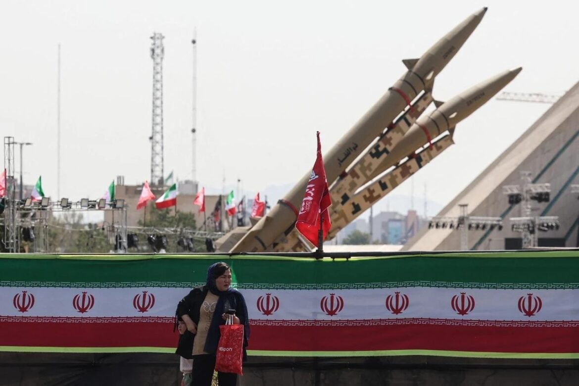 A woman standing near a barrier with Iranian flags, in front of large missiles on display at a public event.