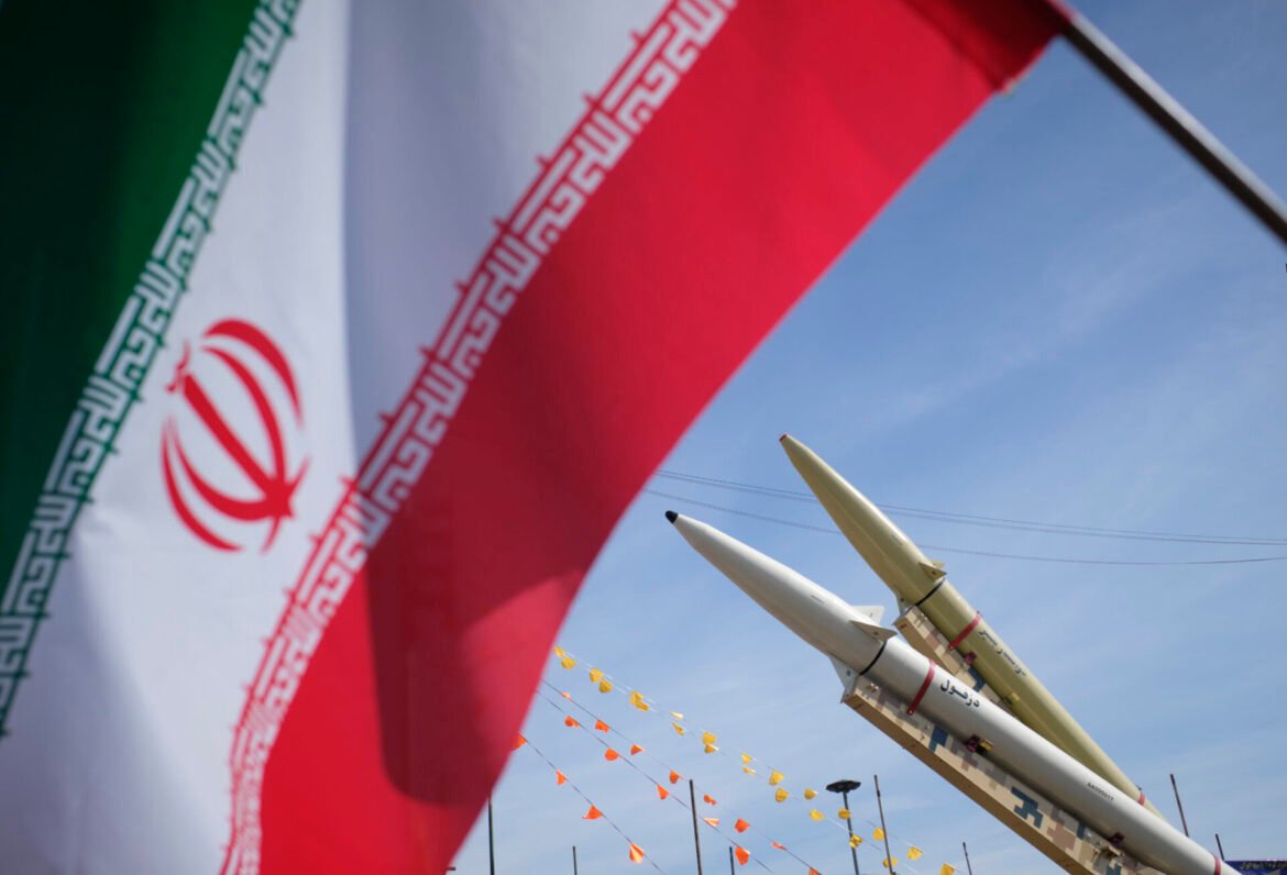 Iranian flag waving with missiles on display against a blue sky.