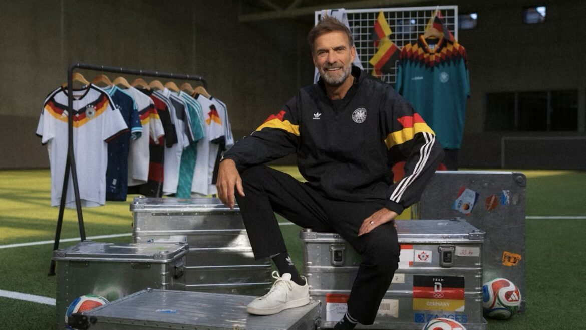 Man in Germany national team gear sits on metal equipment cases in a soccer training area, with jerseys on a rack behind him.