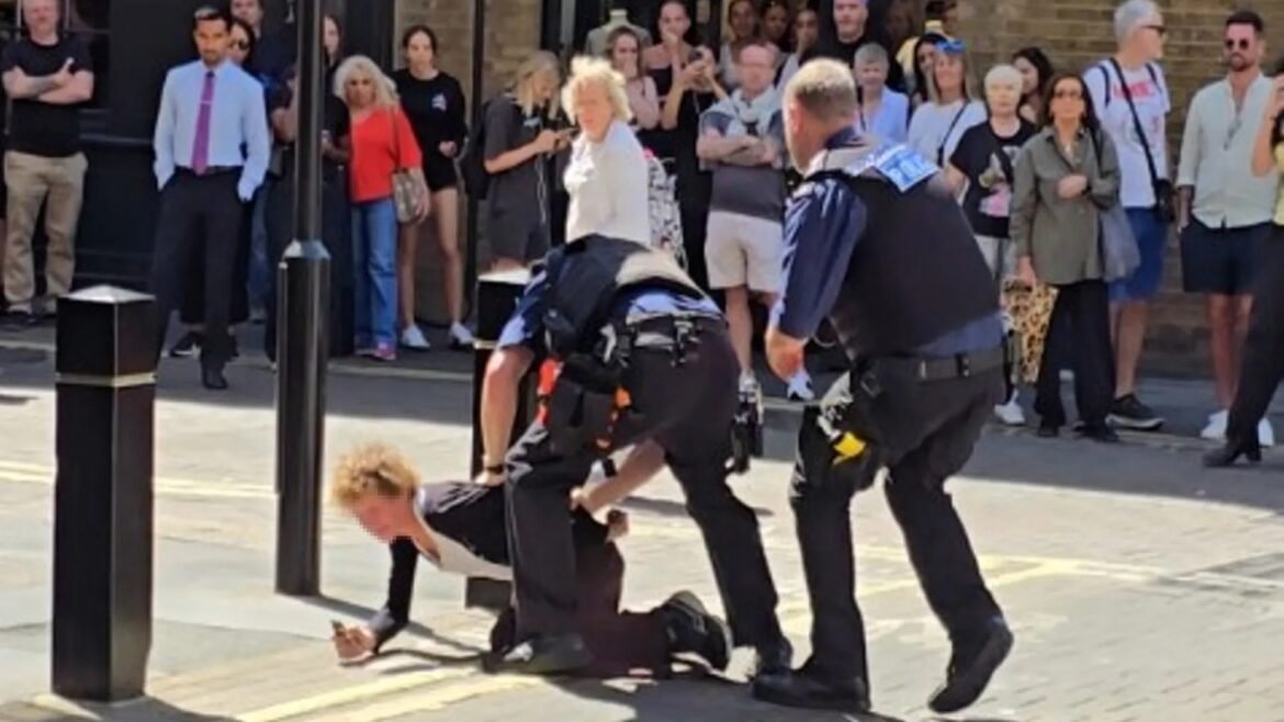 Two uniformed officers restrain a man on the ground while a crowd watches from the sidewalk nearby.