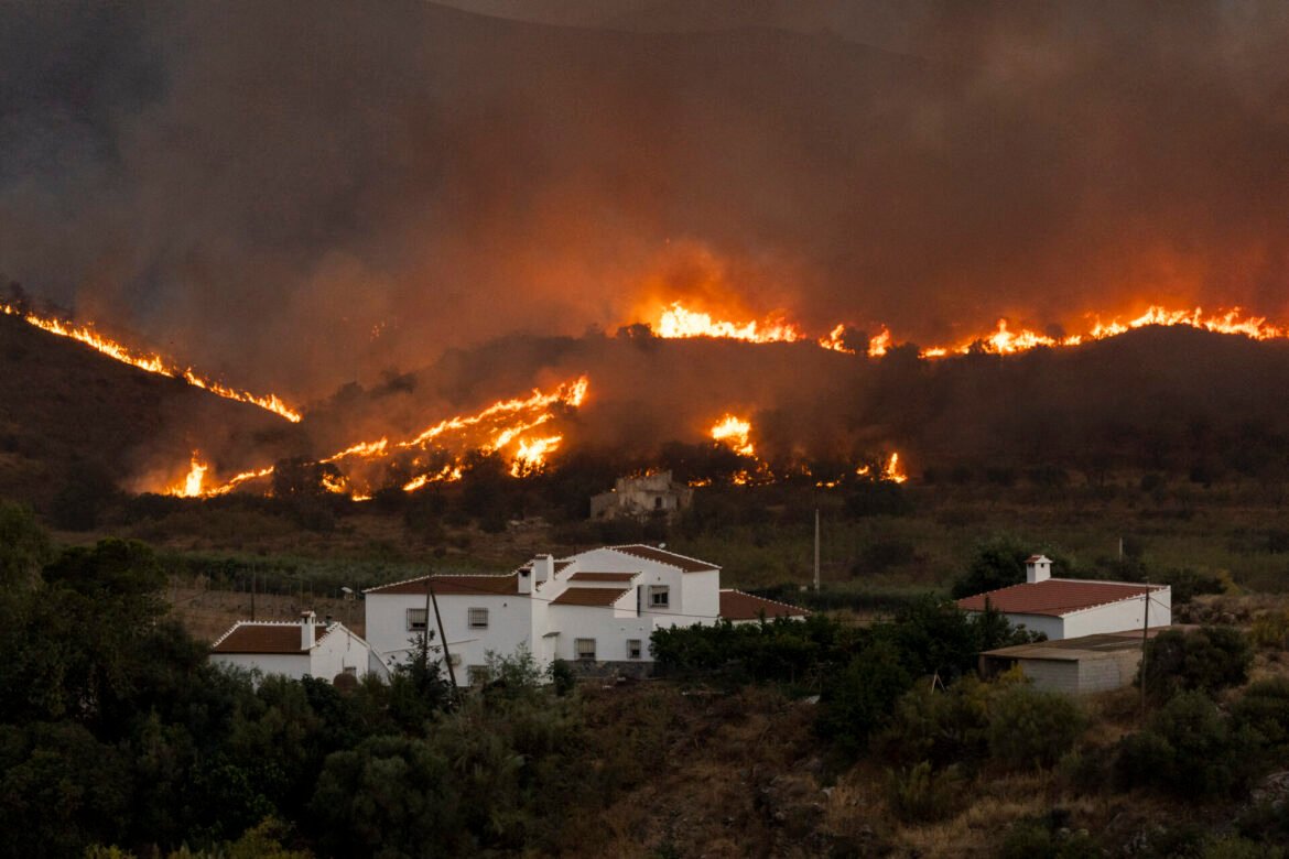 A hillside on fire at dusk with flames along the ridge, small white houses in the foreground and dark smoke filling the sky.