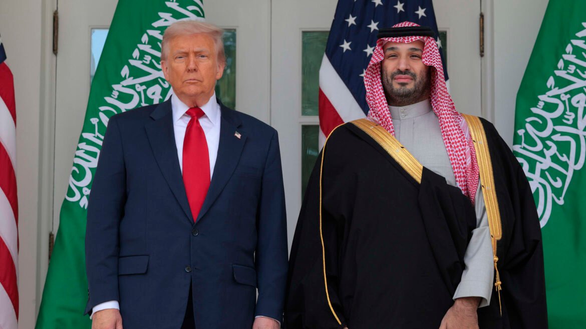 Two men stand side by side in front of flags; one in a navy suit and red tie, the other in traditional Saudi attire with a red-and-white keffiyeh.