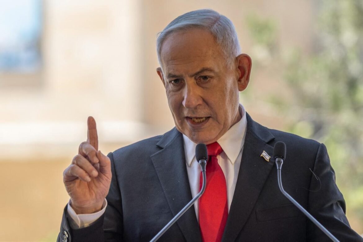 Man in a dark suit and red tie speaks at a podium with two microphones, raising one finger as he gestures while delivering a speech.