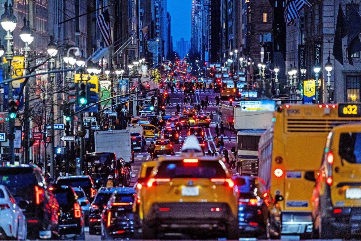 Bustling city street at dusk with yellow taxis, buses, cars, and pedestrians amid illuminated storefronts and streetlamps