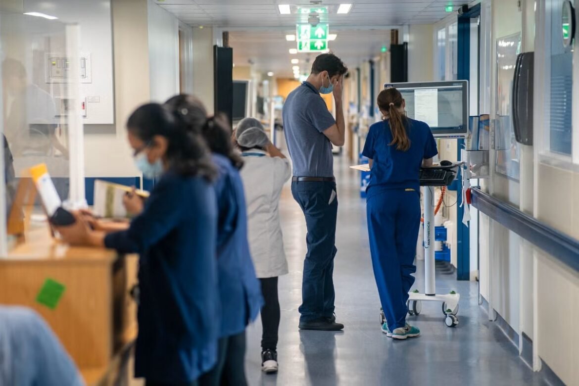Hospital corridor with medical staff; a nurse at a computer cart and a man in blue standing nearby, holding his head in distress.
