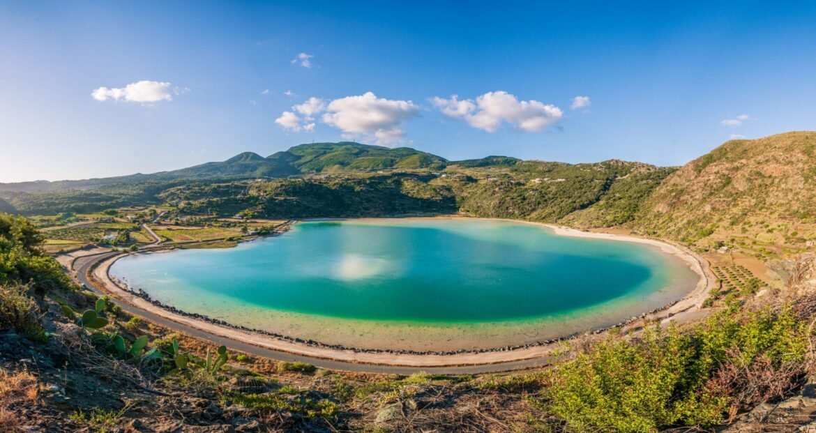 Turquoise circular lagoon bordered by a dirt road and rugged hills under a blue sky with scattered clouds.