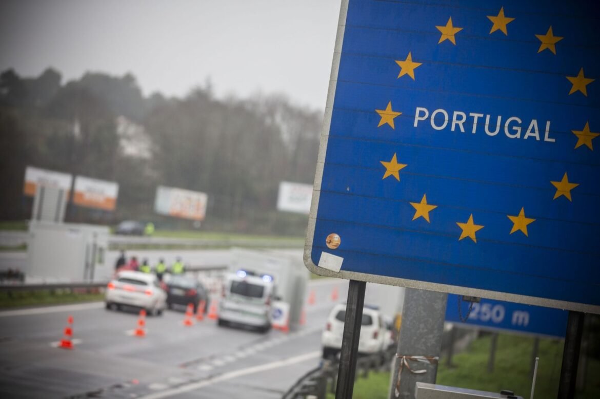 Blue sign with yellow EU stars and the word 'PORTUGAL' at a road border checkpoint, with cones and police/guarded area in the background.