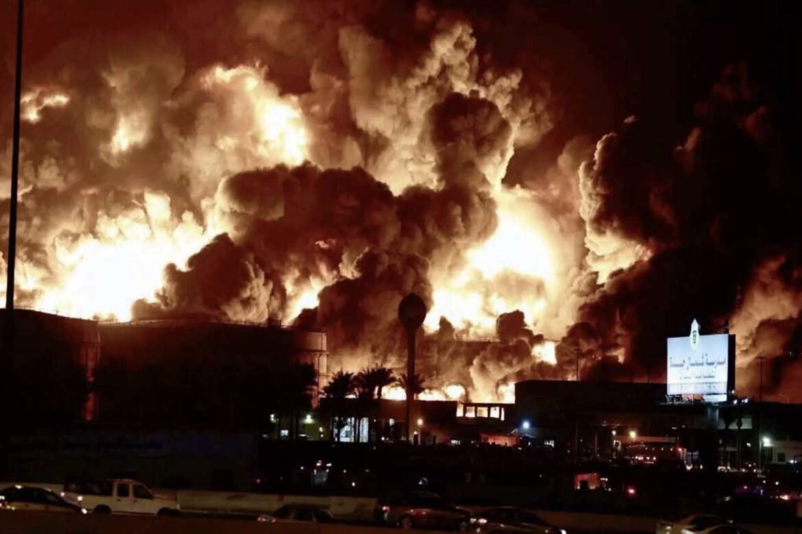 Nighttime scene of a massive industrial fire with towering flames and thick smoke over buildings, emergency lights in the foreground.