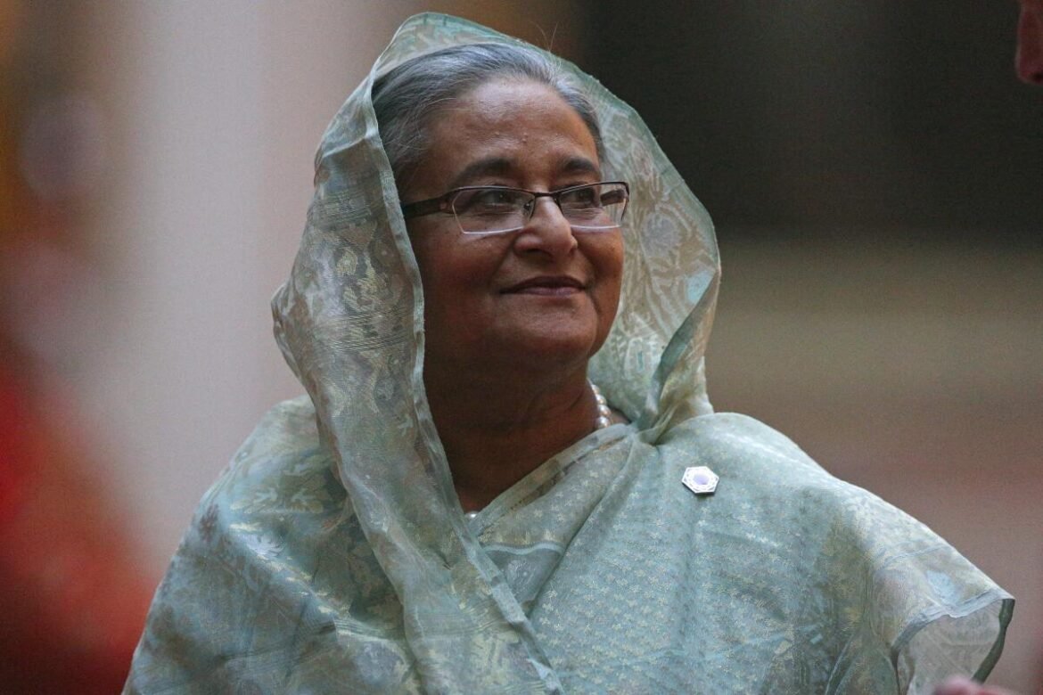 Older woman in a light blue-green patterned sari with head covering, glasses, and a pearl necklace, looking to the side with a gentle smile.