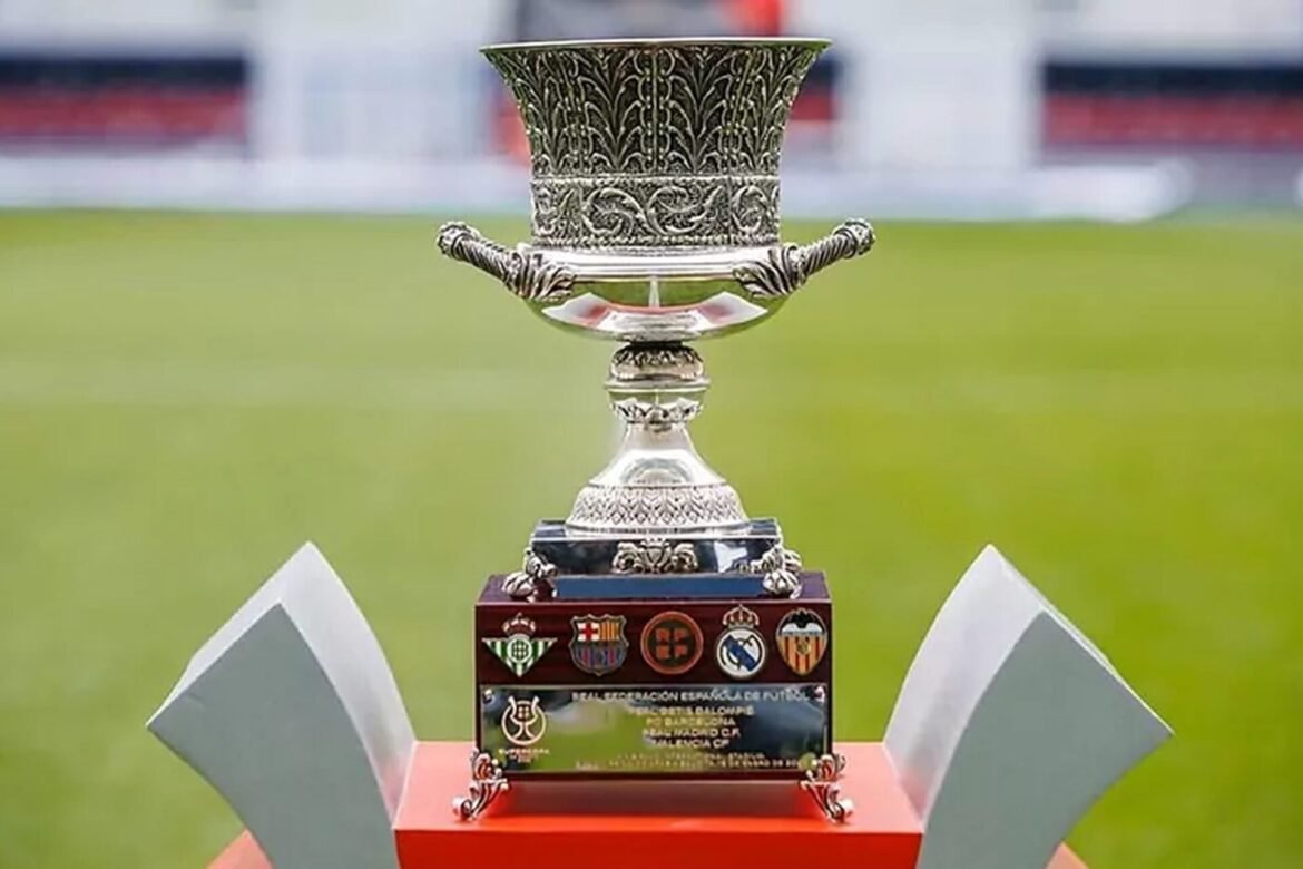 Ornate silver trophy on a red pedestal with football club logos on the plaque, blurred green field background.
