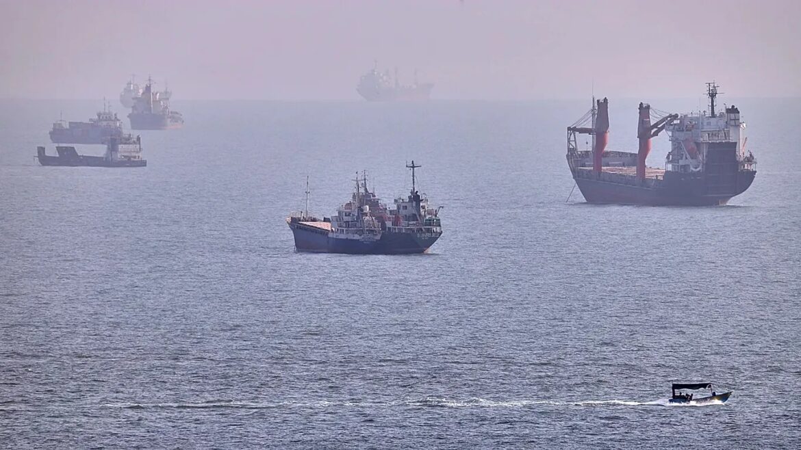 Several cargo ships spread across a calm sea with a small motorboat in the foreground under hazy skies, suggesting a busy harbor scene.