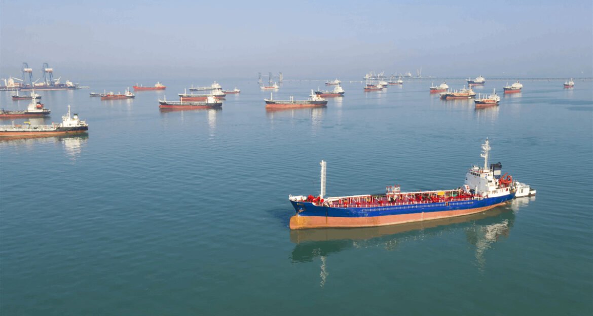A blue cargo ship with a red deck sailing in a calm harbor, dozens of other ships anchored in the distance with port cranes on the horizon.