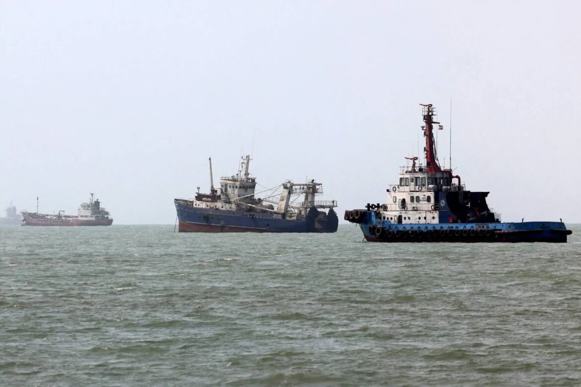 Several large ships anchored on a calm sea; a blue and white tugboat is closest on the right, with two other vessels fading into the hazy horizon.