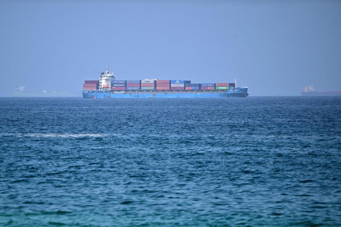 Large blue cargo ship with stacked colorful shipping containers on calm sea, distant vessels on horizon, hazy sky