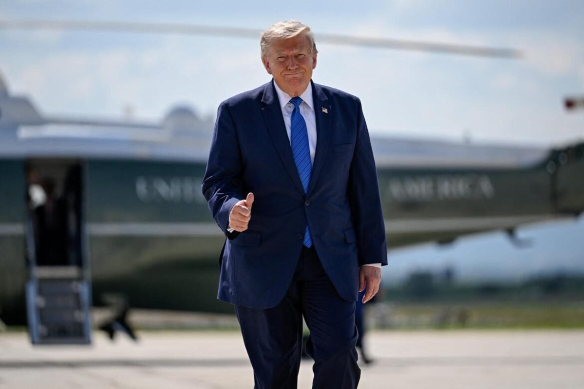 A man in a dark blue suit gives a thumbs-up while walking on a tarmac beside a large aircraft in the background.
