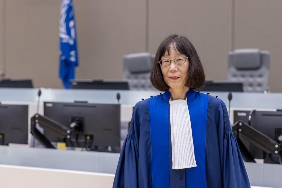 Senior female judge in a blue judicial robe standing in a courtroom with desks, monitors, and a blue flag in the background.
