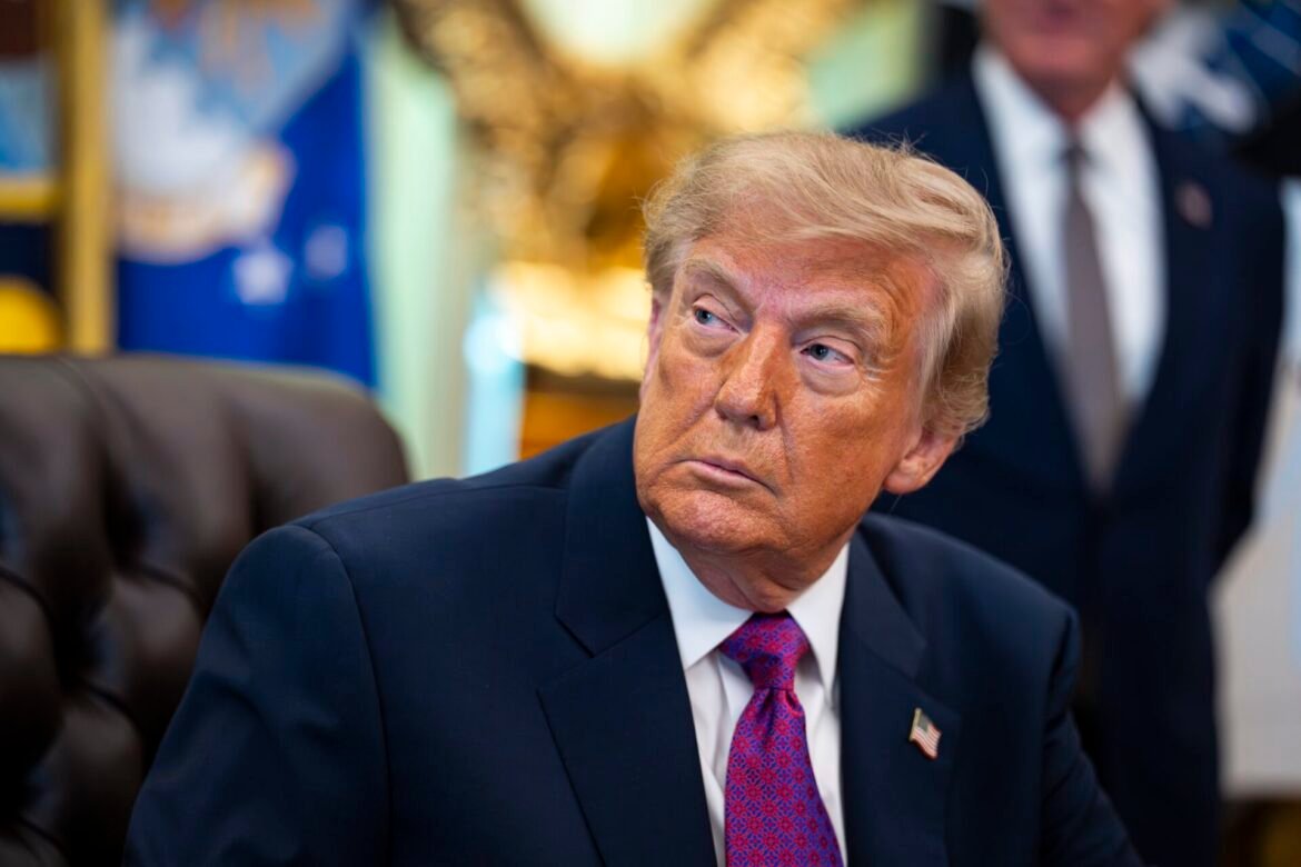 Donald Trump in a dark suit and red tie, seated and looking to his left.