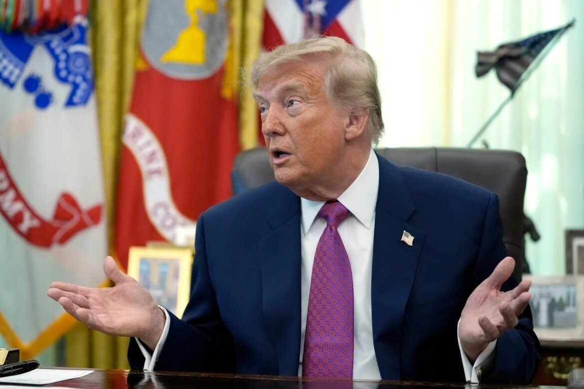 Man in a dark suit and purple tie sits at a desk, hands raised in discussion with flags in the background